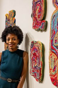 artist standing next to her fiber art hanging on gallery wall